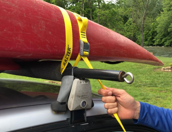 A red kayak secured on top of a vehicle using yellow cam straps with a green landscape in the background.