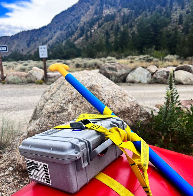A dry box and paddle secured to the back of a red inflatable raft with a yellow cam strap. There is a road and mountains in the background